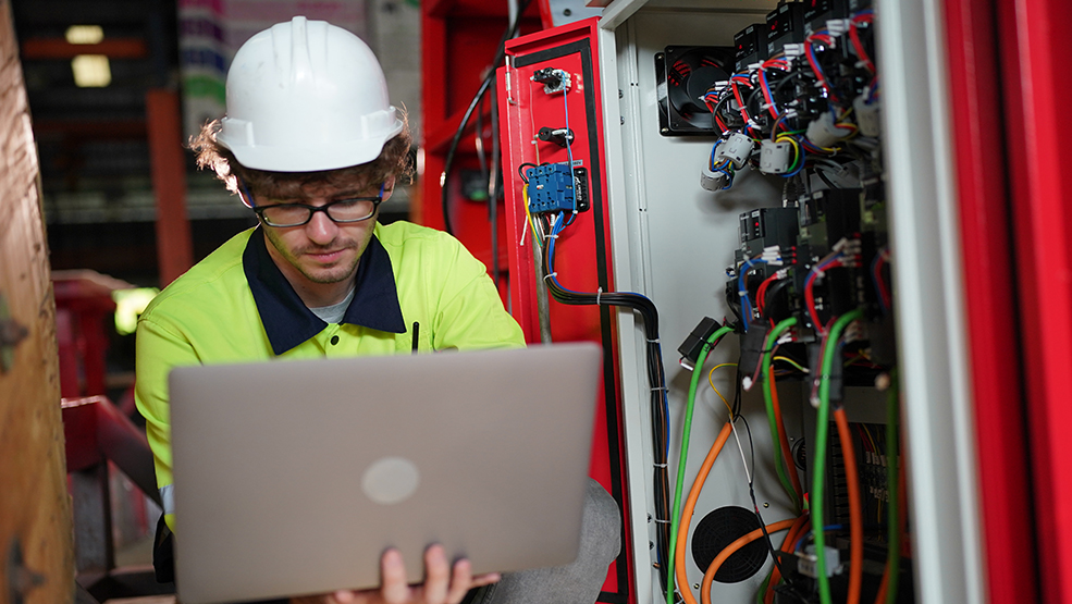 Technician in safety gear using laptop to monitor industrial electrical control panel on-site