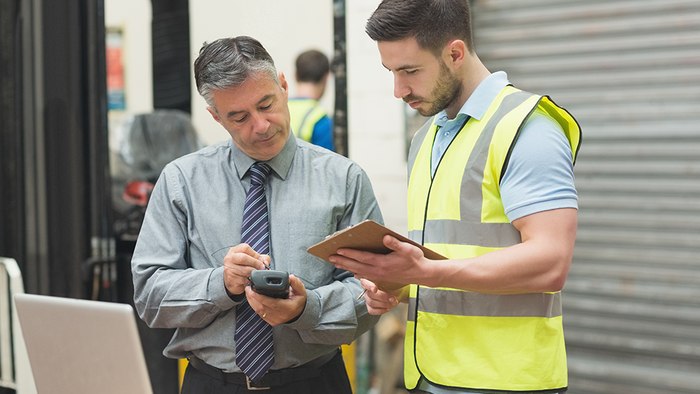 Warehouse manager reviewing handheld scanner data with worker holding clipboard in logistics facility