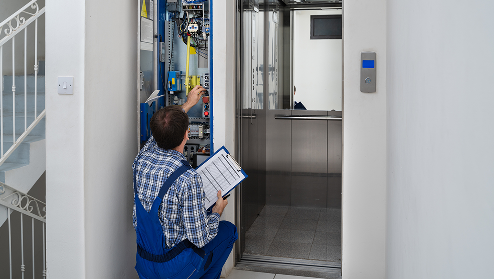 Elevator technician reviewing inspection checklist while examining electrical control panel beside open lift