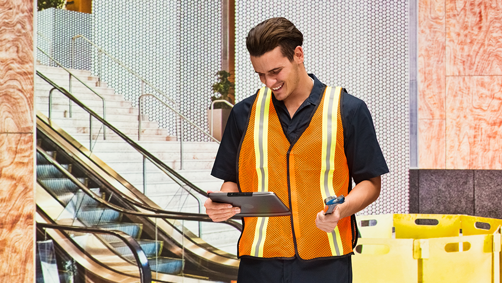 Field service technician in safety vest reviewing tablet and handheld device near escalator at commercial job site