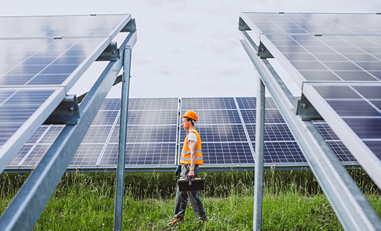 Solar panel technician in safety gear walking through a ground-mounted photovoltaic array at a solar farm