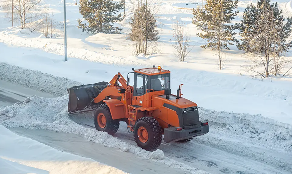 Orange wheel loader clearing snow from a residential road in winter