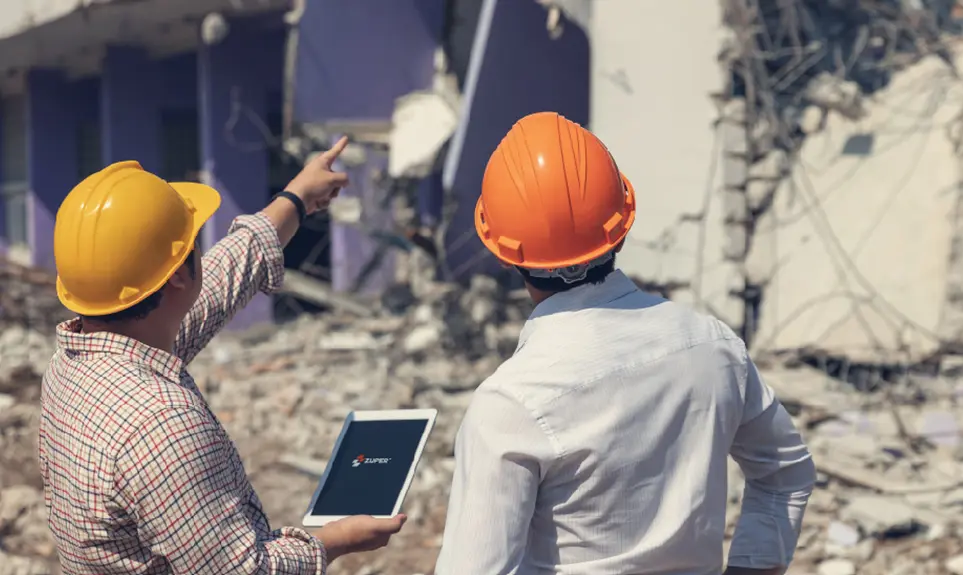 Two construction workers in hard hats review a tablet while assessing building damage at a demolition site