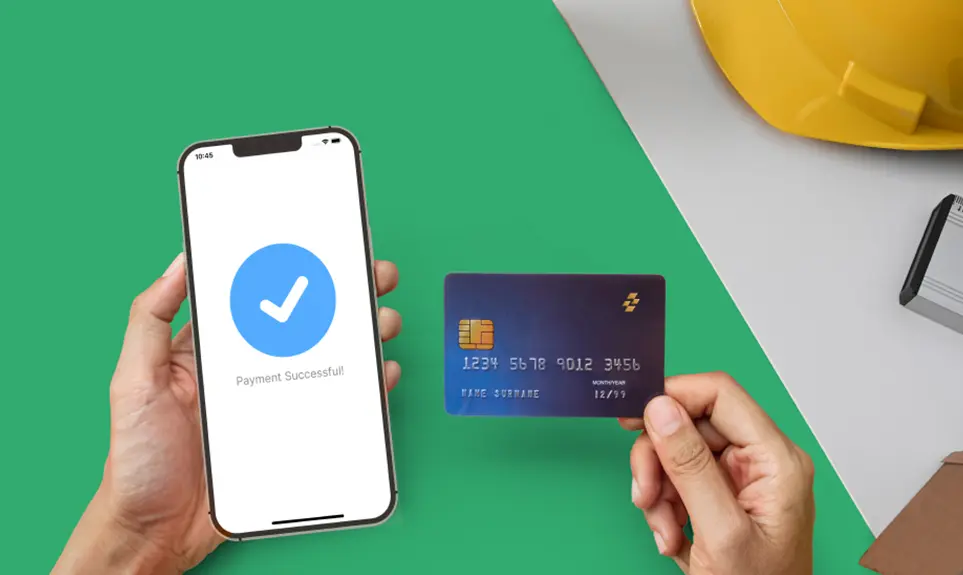 Mobile payment confirmation screen with checkmark beside credit card held over green workspace with hard hat