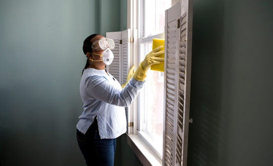 Woman wearing protective mask, goggles, and gloves cleans window shutters with yellow cloth