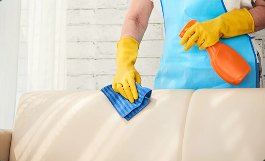 Professional cleaner wiping beige leather couch with blue microfiber cloth and spray bottle