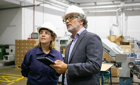 Factory supervisor with tablet discussing operations with manager wearing hard hats on manufacturing floor
