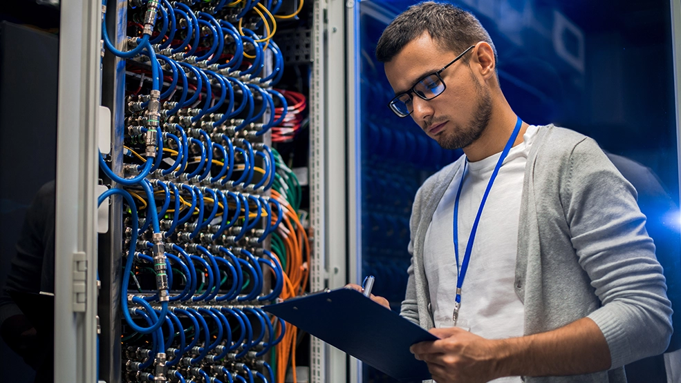 IT technician documenting server rack equipment and network cable connections on clipboard in data center