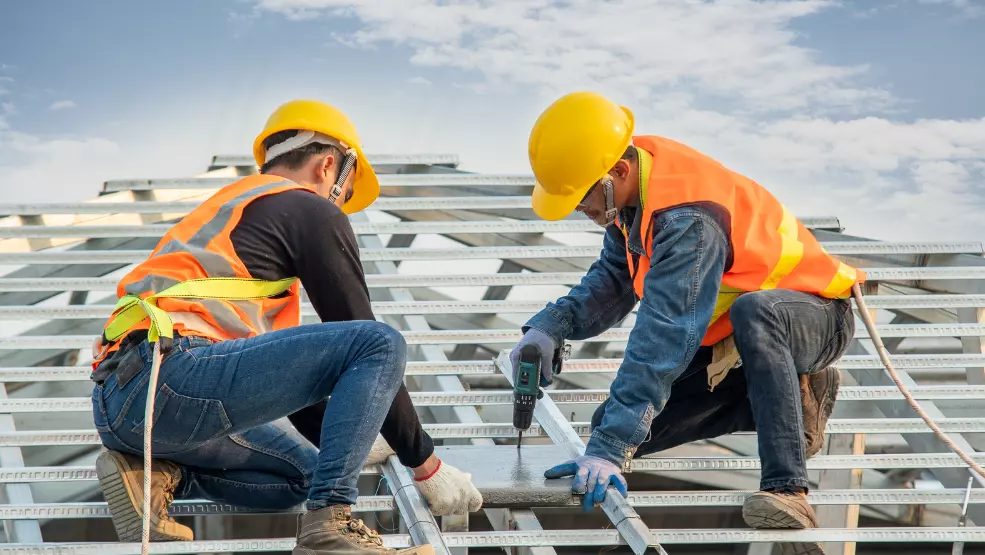 Two roofing contractors in safety gear installing steel roof framing using a power drill