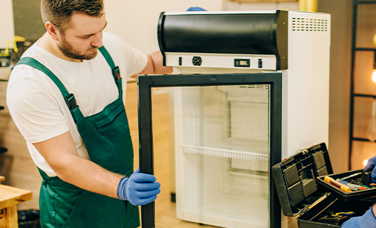 Technician in green overalls and blue gloves repairing a commercial display refrigerator with toolbox nearby