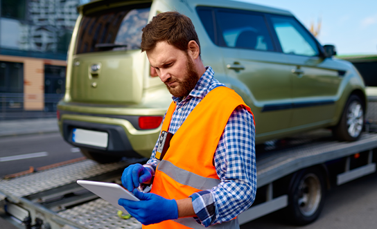 Tow truck operator in safety vest reviewing digital paperwork on tablet while loading vehicle onto flatbed