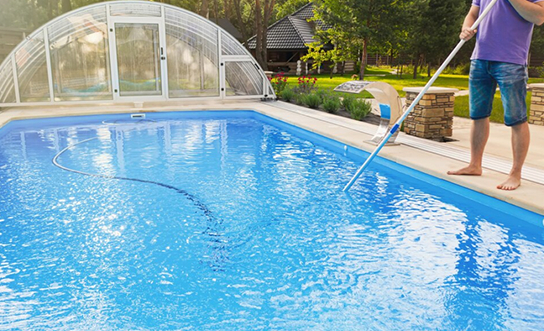 Pool technician using telescopic pole to clean residential swimming pool with retractable enclosure