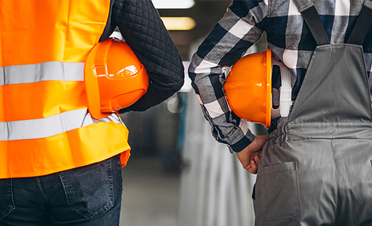 Two construction workers holding orange hard hats at their sides in an industrial setting