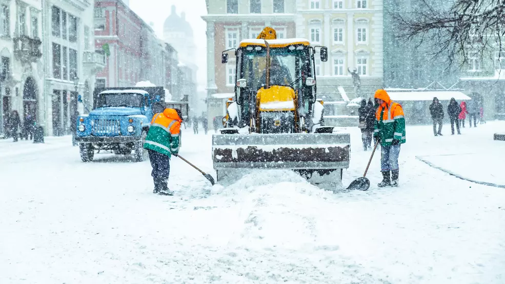 Snow removal crew shoveling alongside a front-loader plow clearing a city street during heavy snowfall