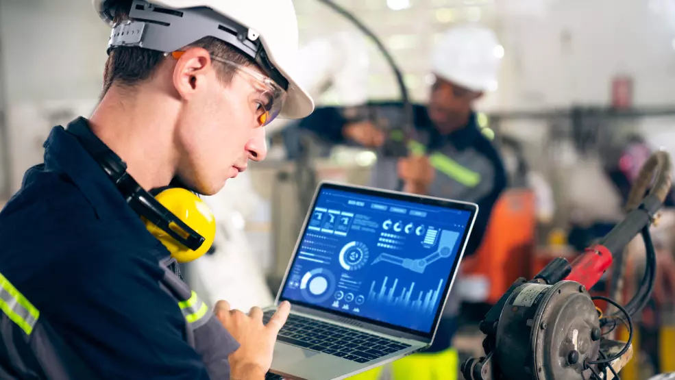 Field service technician in safety gear using laptop to monitor industrial equipment performance data and analytics