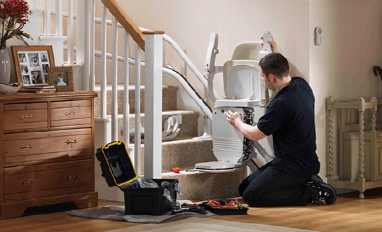 Technician kneeling to repair a stairlift at the base of a carpeted staircase with tools spread on the floor