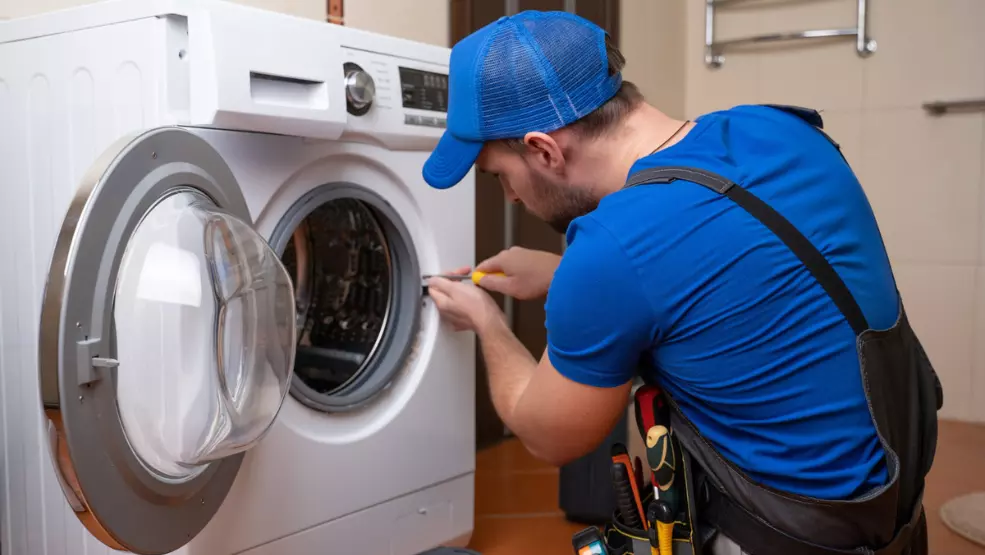 Technician in blue uniform using screwdriver to repair front-load washing machine door seal