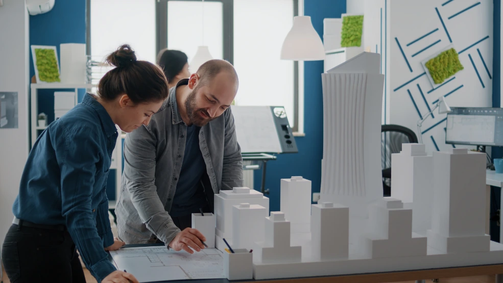 Two architects reviewing blueprints at a desk with a white architectural city model in a modern design studio