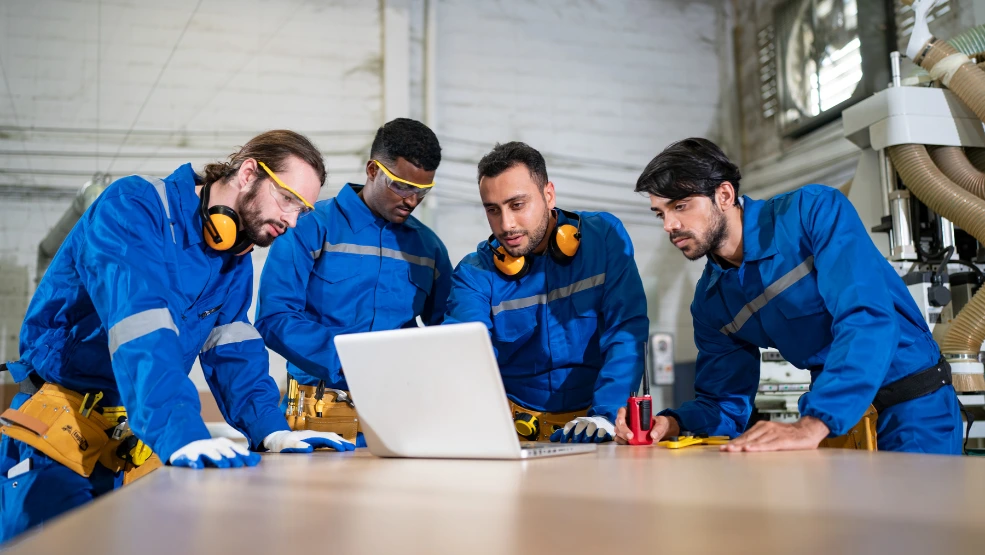 Four technicians in blue coveralls with safety gear reviewing information on a laptop in an industrial workspace