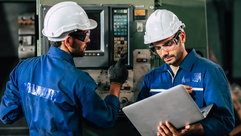 Two technicians in hard hats and safety glasses reviewing data on a laptop near industrial control equipment