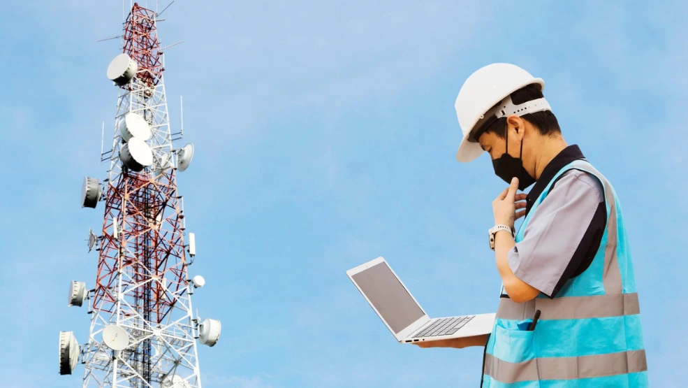 Telecom technician in safety vest and hard hat reviewing data on laptop near cell tower