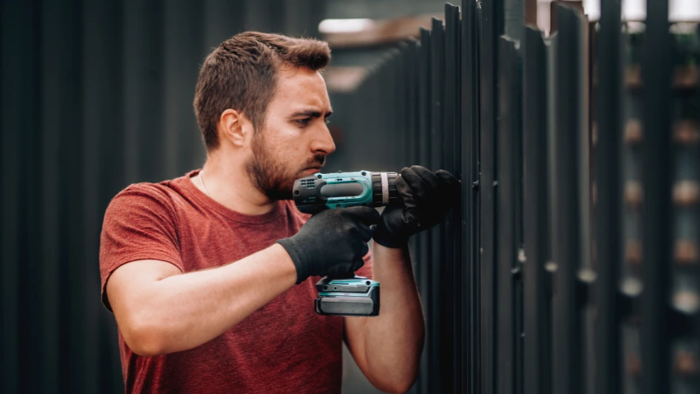Contractor using a cordless drill to install a dark metal fence panel