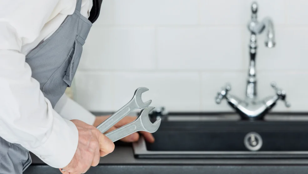 Plumber in gray apron holding two wrenches near kitchen sink faucet ready for repair work