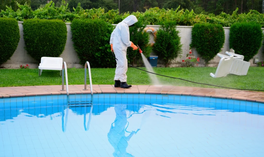 Pool service technician in protective suit pressure washing patio furniture beside a residential swimming pool