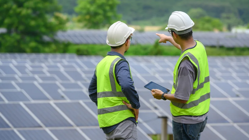 Two solar technicians in safety vests and hard hats reviewing data on a tablet at a solar panel installation site