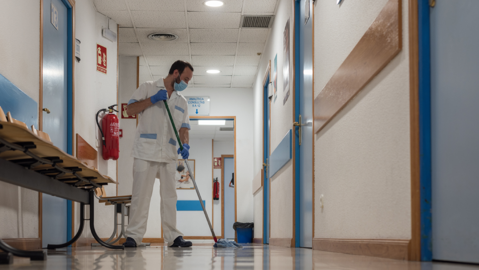 Custodial worker mopping hospital corridor floor while wearing protective mask and gloves