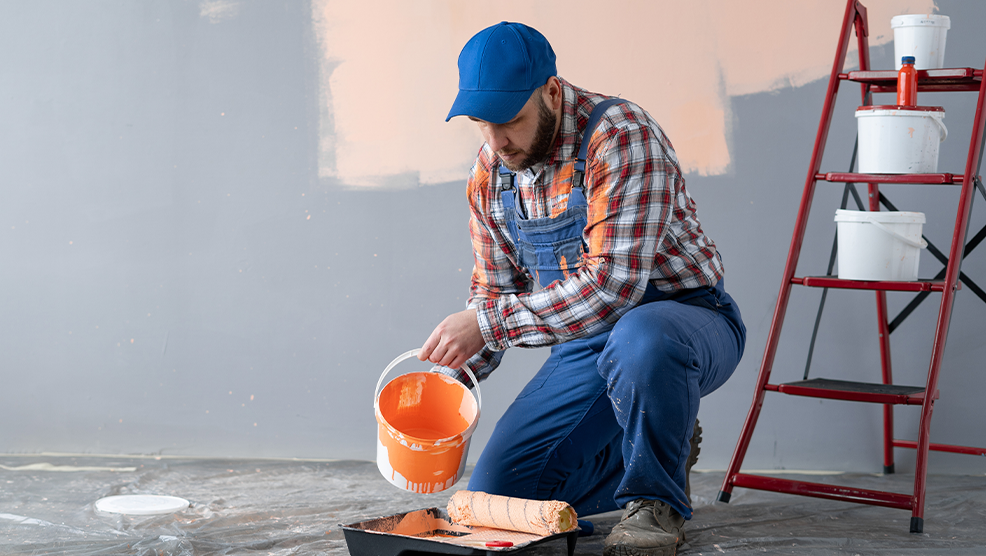 Painter pouring orange paint into a tray while preparing to paint a wall during a home renovation project