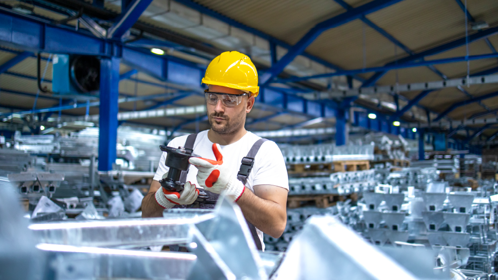 Factory worker in yellow hard hat and safety glasses inspecting a metal component in an industrial manufacturing facility