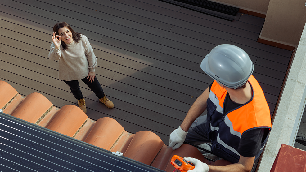 Smiling homeowner gives okay sign to roofing technician installing solar panels on terra cotta tile roof