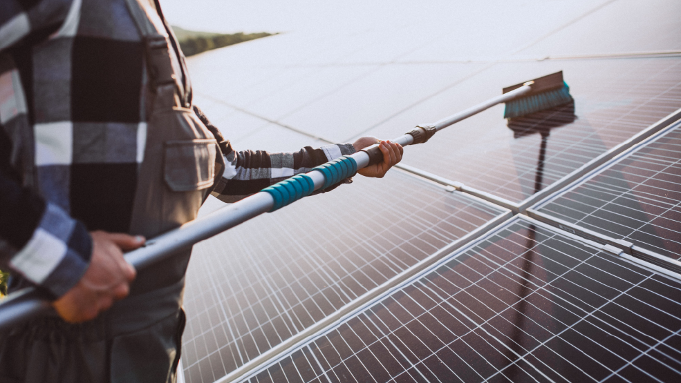 Technician cleaning solar panels with an extendable brush at a ground-mounted solar installation