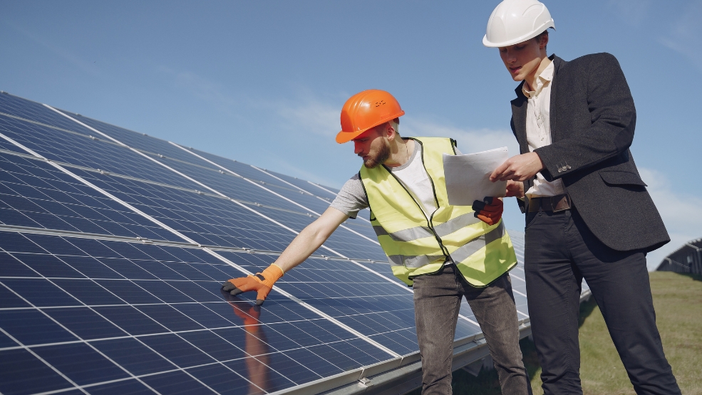 Solar technician in safety vest inspecting panels while supervisor reviews documentation at ground-mounted installation site