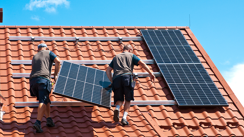 Two technicians installing solar panels on a terracotta tile roof on a sunny day