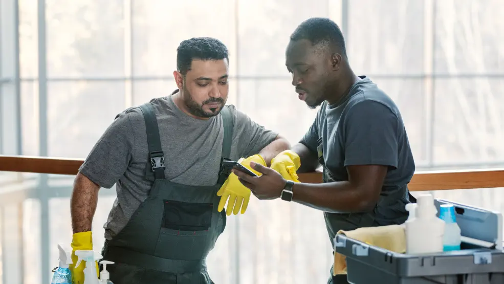 Two cleaning professionals in yellow gloves reviewing a mobile device while working with cleaning supplies