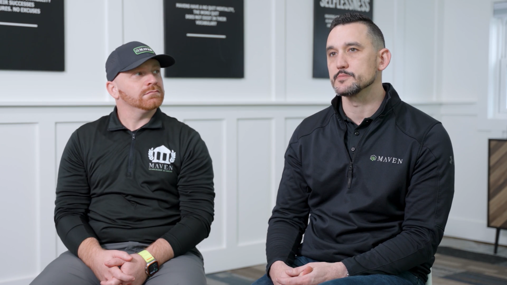 Two men in black Maven-branded quarter-zip shirts seated during an interview in an office with motivational posters