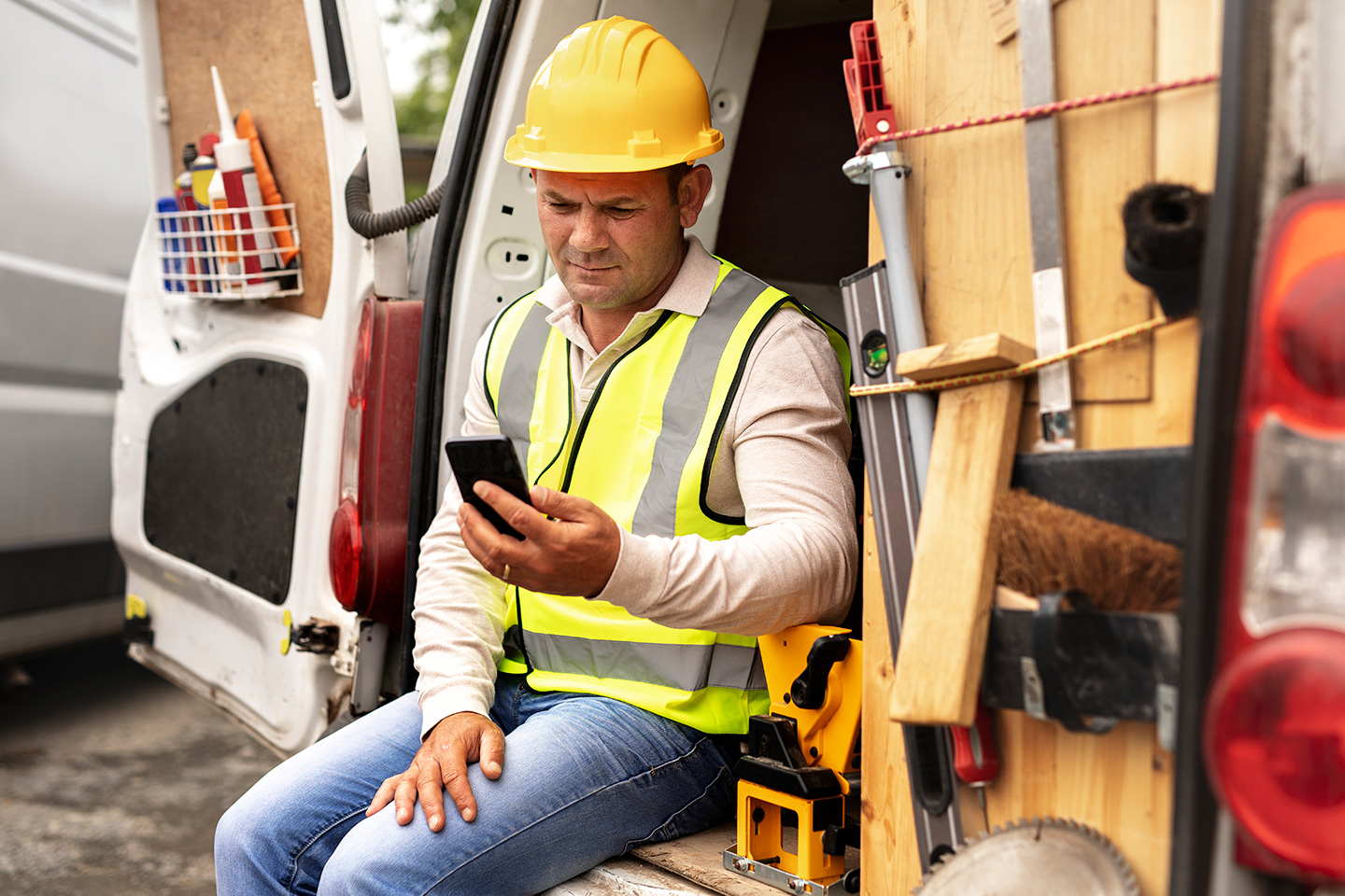 Contractor in hard hat and safety vest checking smartphone while seated in work van with tools