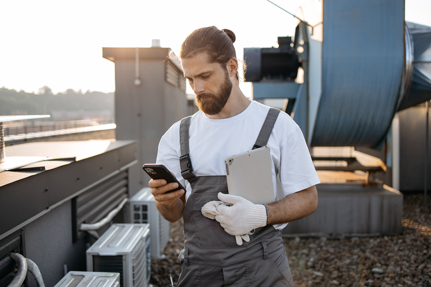 HVAC technician in work overalls checking smartphone while holding tablet on commercial building rooftop near ventilation equipment