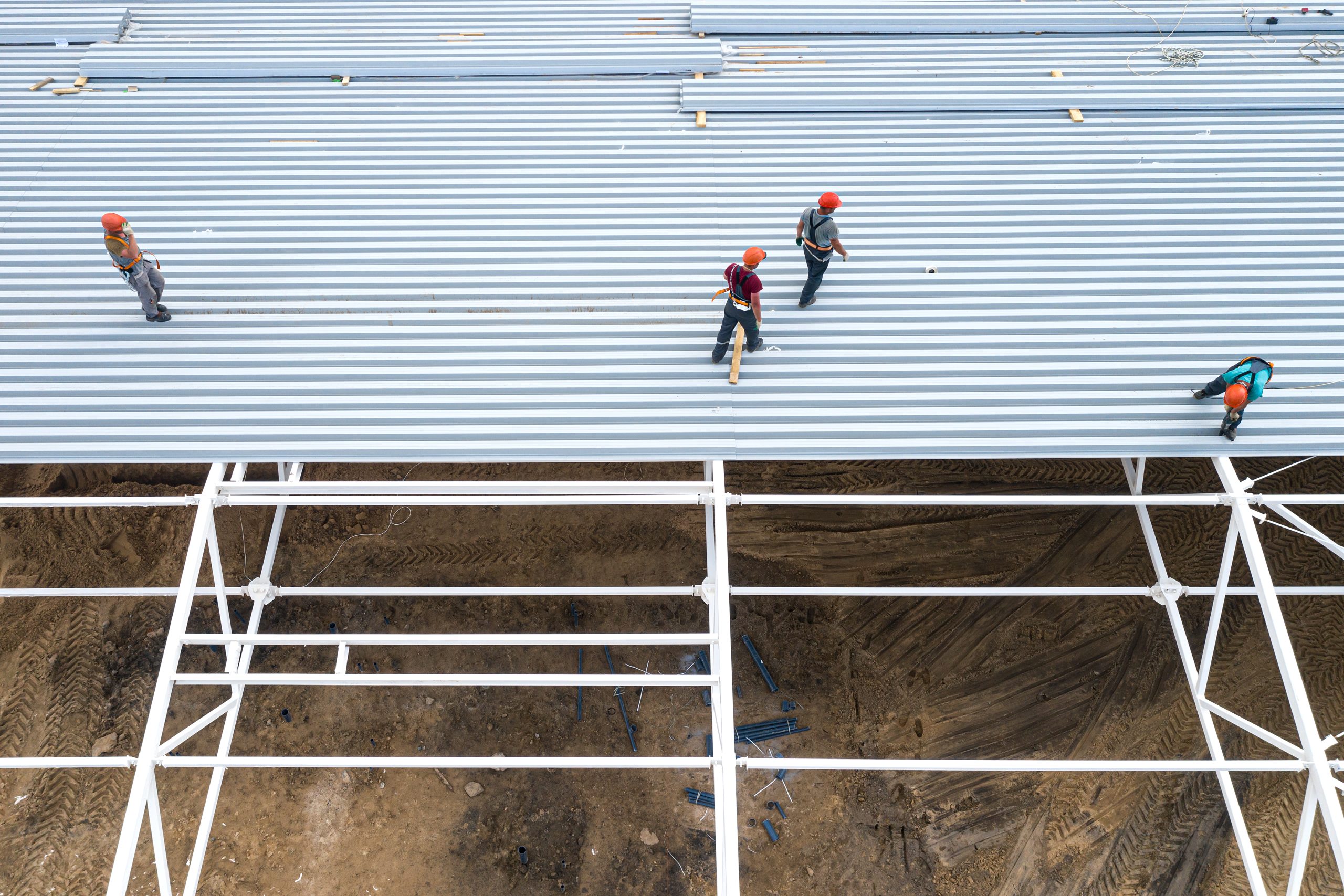 Construction workers in orange hard hats installing corrugated metal roofing panels on a commercial building steel frame