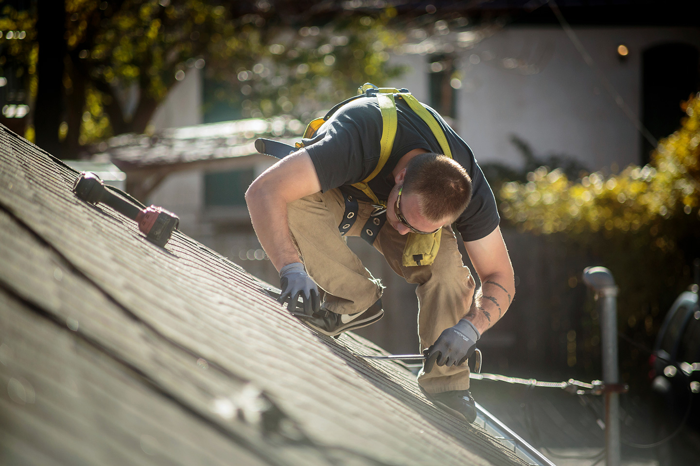 Roofer in safety harness securing metal rail on shingled roof during solar panel installation