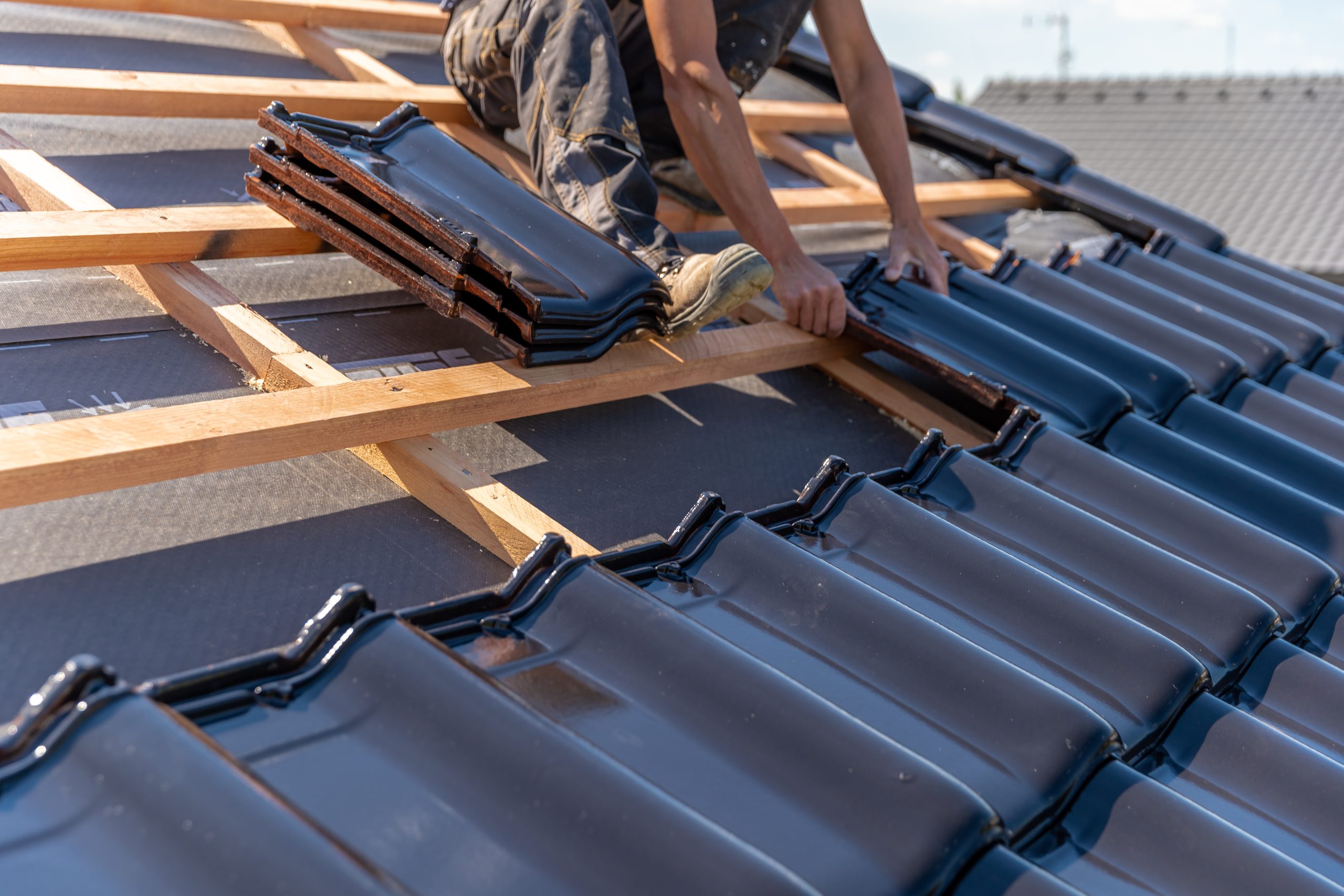Roofer installing dark ceramic roof tiles on wooden battens during residential roof construction