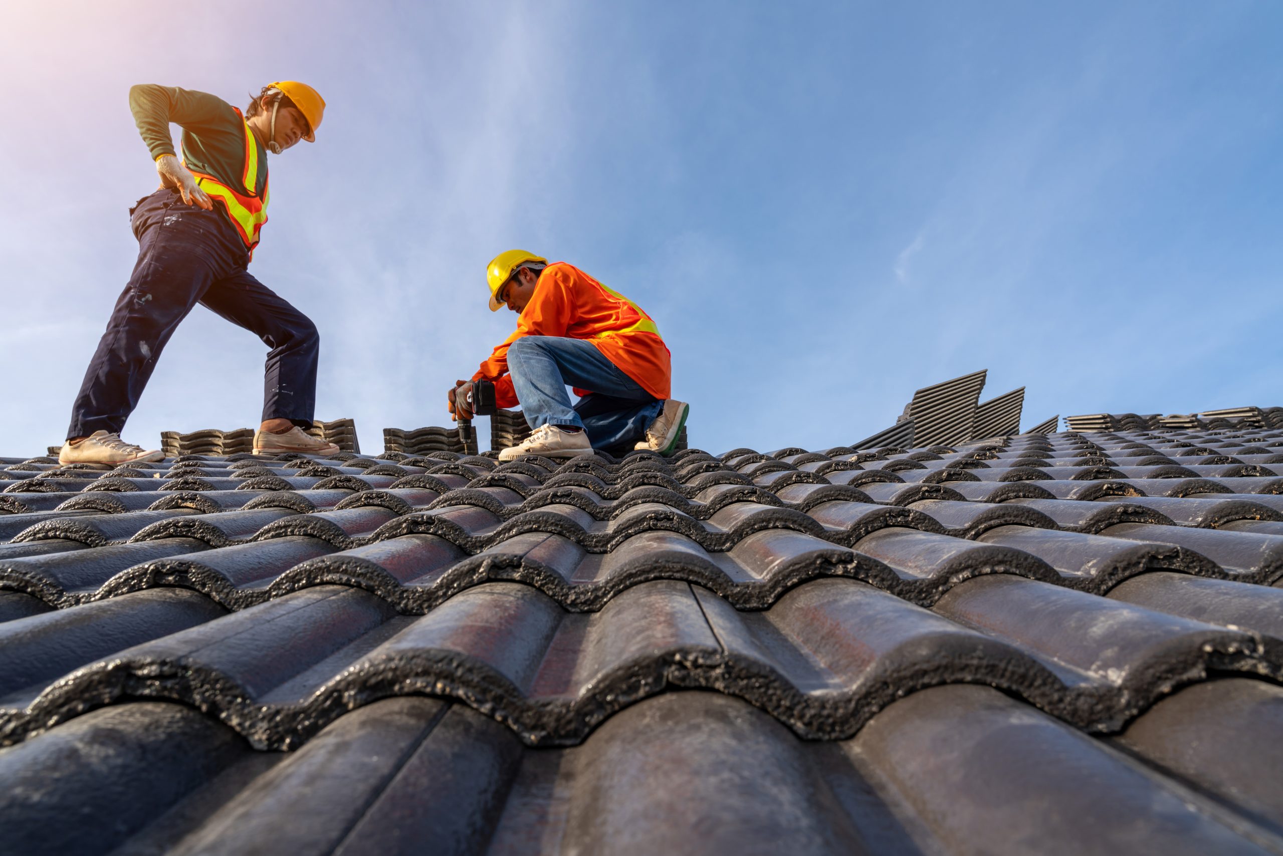 Two roofers in safety vests and hard hats installing dark clay tiles on a residential roof
