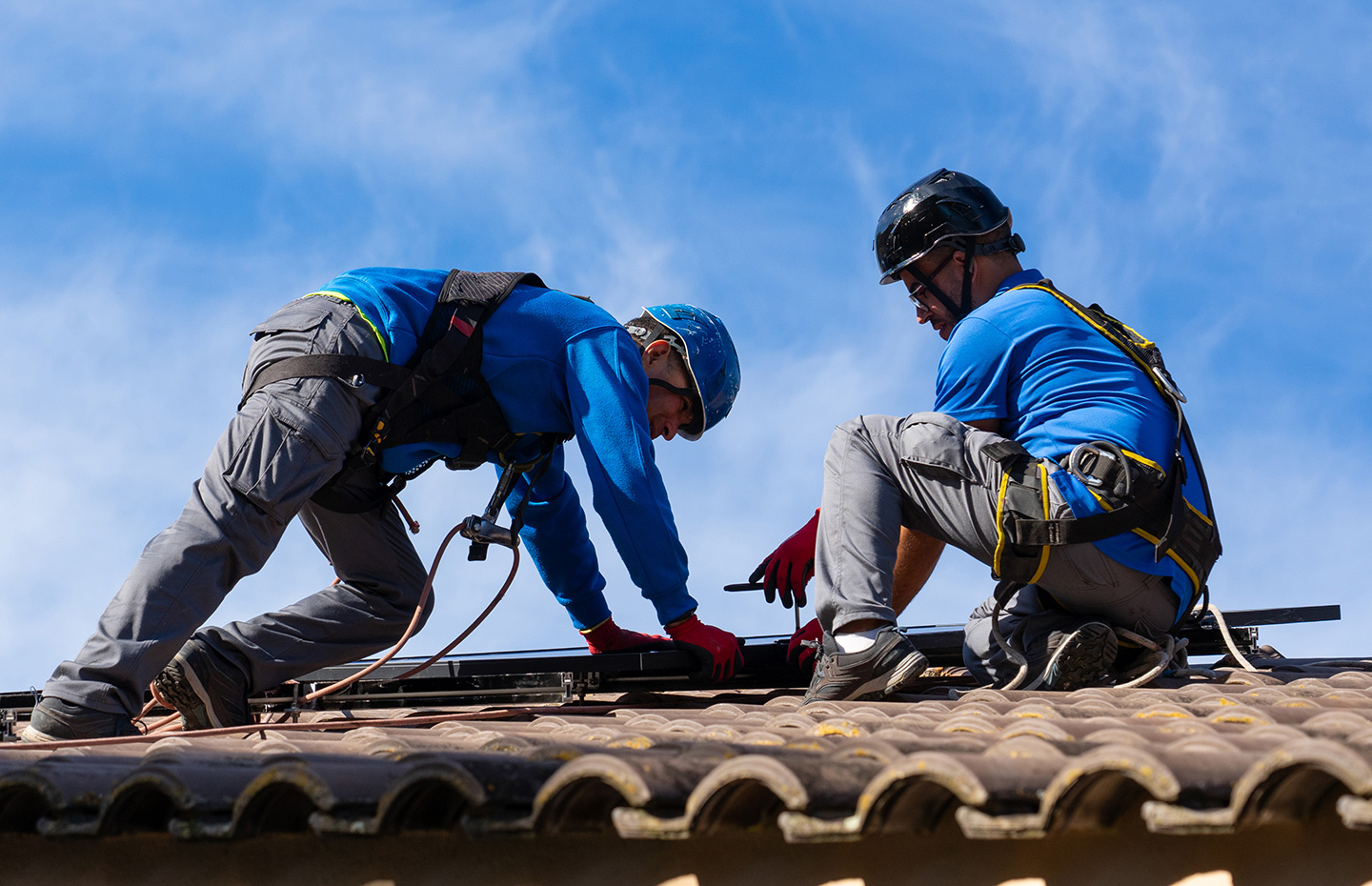 Two roofing technicians in safety harnesses installing solar panel mounting rails on a clay tile roof