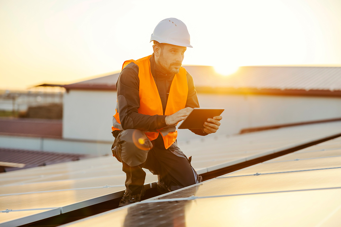 Roofing technician in safety vest and hard hat inspecting solar panel installation with tablet on commercial roof
