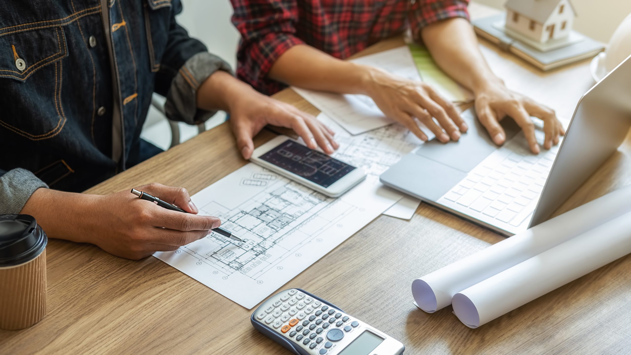 Two professionals reviewing architectural blueprints at a desk with laptop, calculator, and coffee while planning a project
