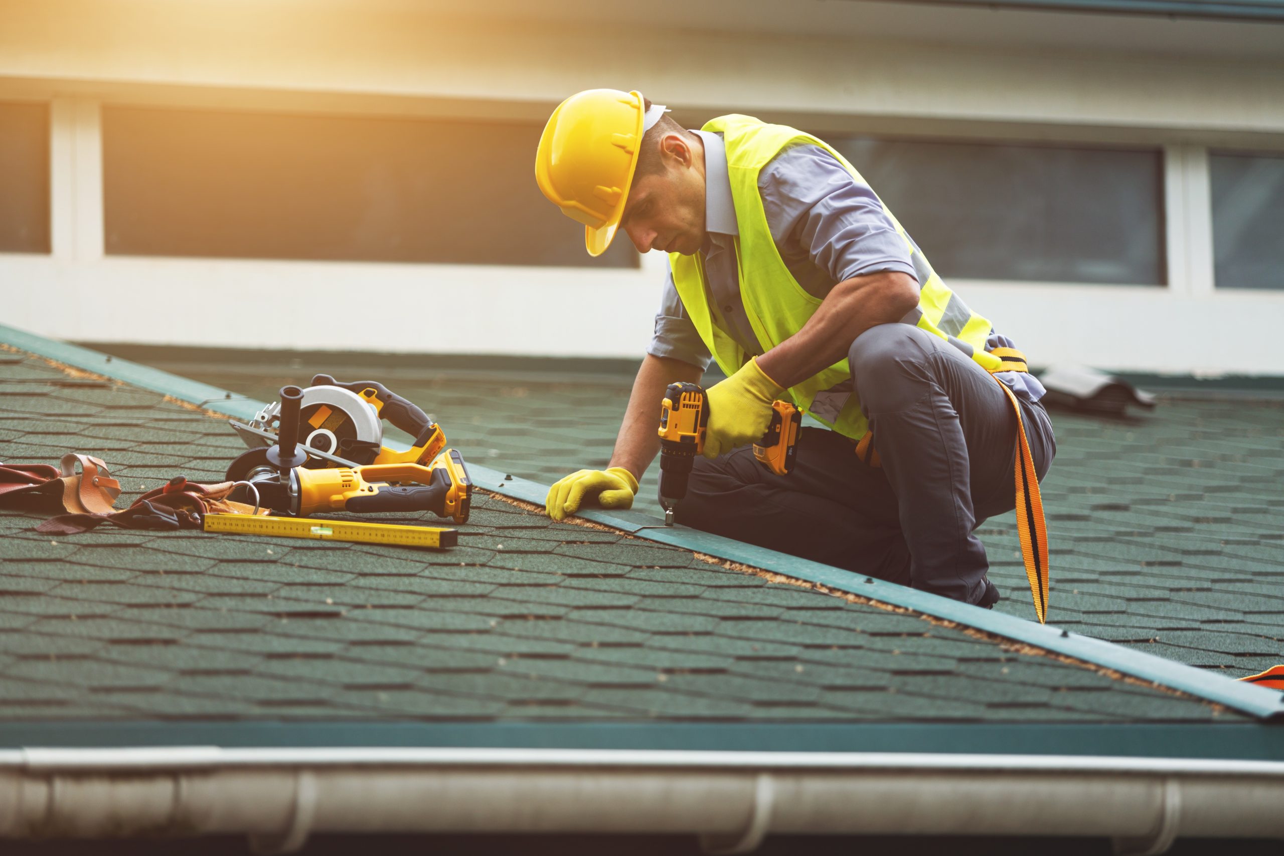 Roofer in safety gear using a cordless drill to install asphalt shingles on a residential roof