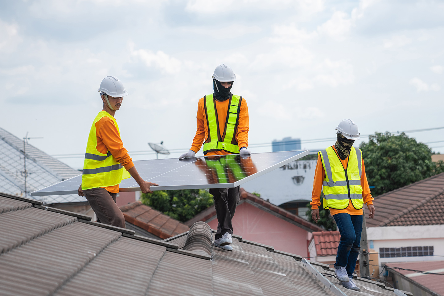 Three workers in safety vests and hard hats carry a solar panel across a residential rooftop for installation